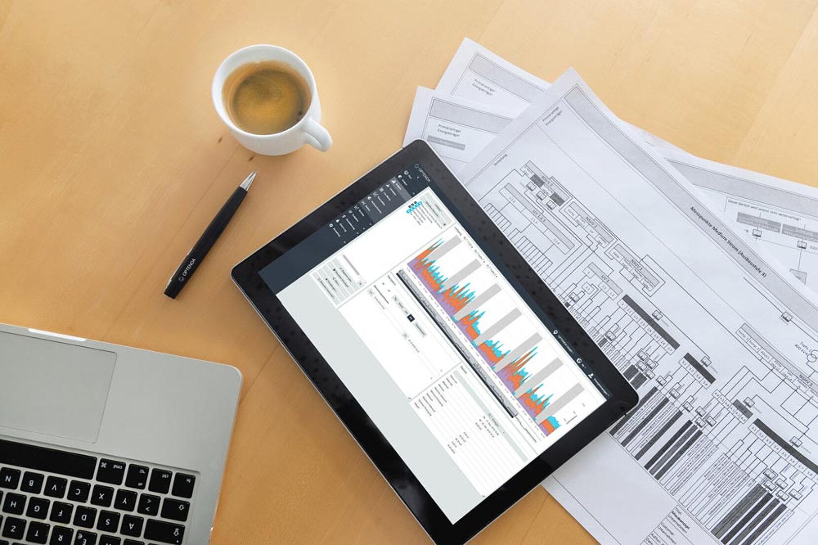 Tablet mit Energy Monitor Software von OPTENDA mit abgebildetem Lastgang neben Kaffeetasse und Stift auf ausgedrucktem Messkonzept